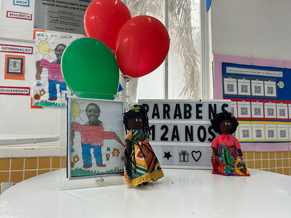 Na imagem podemos ver uma mesa comemorativa onde se encontra a foto da homenageada que dá nome a escola e um pequeno letreiro com os direzeres "parabéns 12 anos" além de balões de festa nas cores vermelha e verde e duas pequenas bonequinhas vestidas com roupas de estampas que remetem a cultura negra.