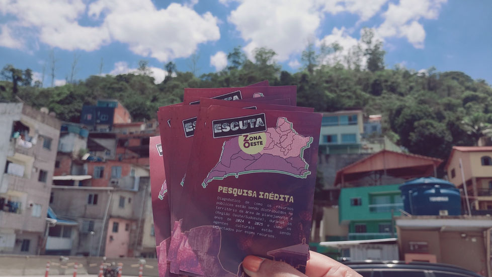 Na foto podemos ver uma mão segurando panfletos do projeto e de fundo a vista de uma comunidade