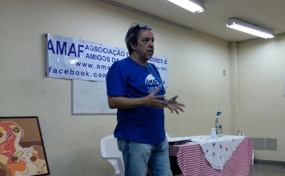 Na imagem podemos ver um homem branco, com cabelo pretos lisos, ele veste uma blusa azul com o símbolo da Associação de Moradores do bairro da Freguesia, e calça, jeans, seus óculos estão apoiados em sua cabeça, como quem levanta os óculos após uma leitura. O homem está de pé em uma sala de paredes amarelo claro e pode-se ver uma faixa com o nome AMAF pendurada na parede atrás do homem.