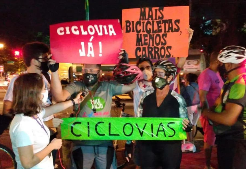 Na imagem podemos ver algumas pessoas segurando faixas com palavras de ordem a respeito do pedido de mais ciclovias na região de Jacarepaguá. Uma repórter está entrevistando algumas das pessoas que usam trajes específicos para a prática do ciclismo.