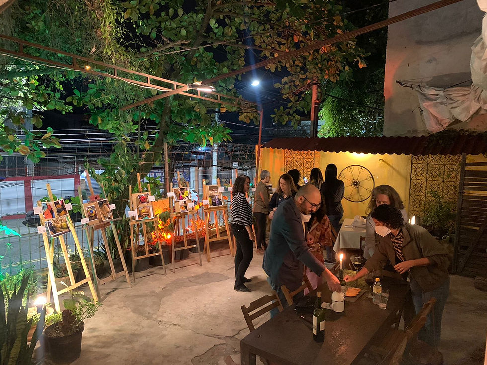 Na imagem podemos ver uma área do restaurante onde está sendo realizada uma exposição de arte com diversos quadros, além disso podemos ver pessoas conversando e arrumando elementos e objetos em uma mesa de madeira.
