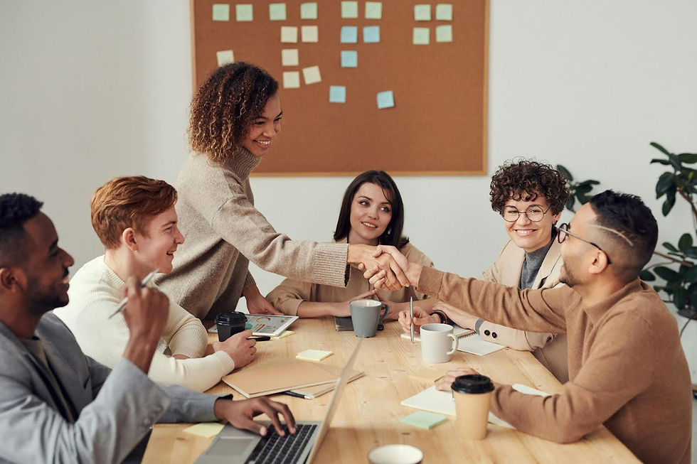 Group of six people in a meeting room. Two shake hands at a wooden table with laptops and coffee. Sticky notes on corkboard in background.