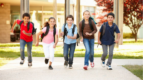 Five smiling children with backpacks run on a school path. The background has green grass, trees with red leaves, and a building.