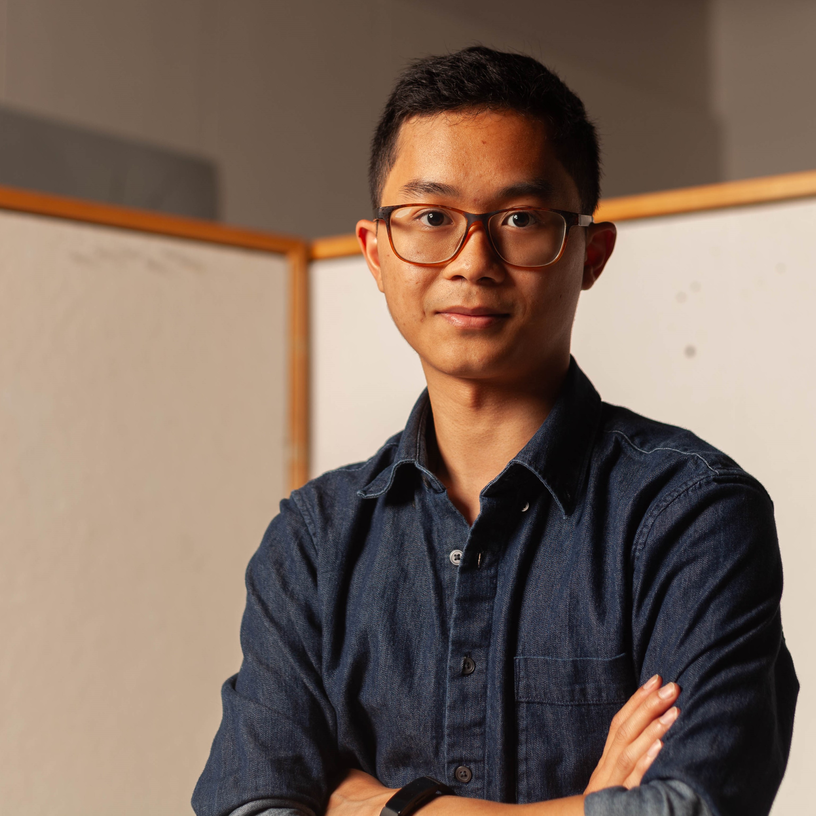 Alex, dressed in a denim shirt and crossing his arms, poses in front of a white background