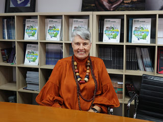 Debra Roberts sits at a wooden desk in front of bookshelves filled with books. She has short gray hair and wears an orange silk blouse and a large bead necklace.