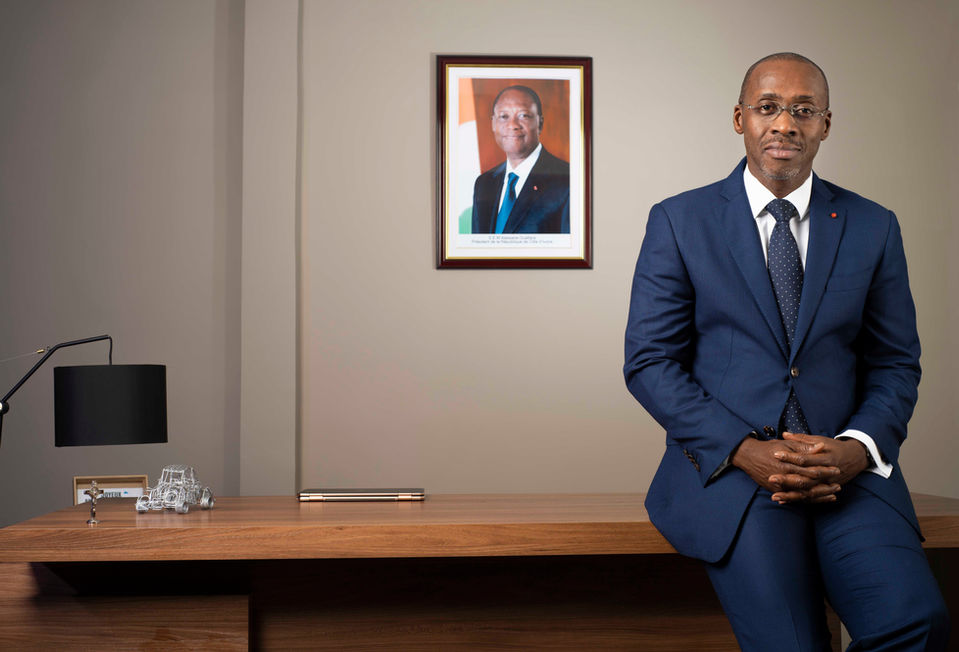 N'ZI Assamoua Désiré Directeur General ARTI assis sur le bord de son bureau en bois en veste et cravate bleu avec en fond la photo officielle du President de la Republique de Cote d'Ivoire Alassane Ouattara sur le mur et sur le bureau un ordinateur portable fermé, un petit crucifix, une petite voiture en fer