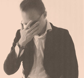 A black and white photograph of a person in a dark blazer and open white shirt, hand raised to cover their face, against a plain white background.