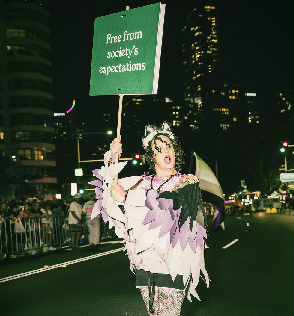 Ashley Olive is marching in the Sydney Mardi Gras, wearing a costume with paper wings and their mouth open as though they are calling something out loud.
