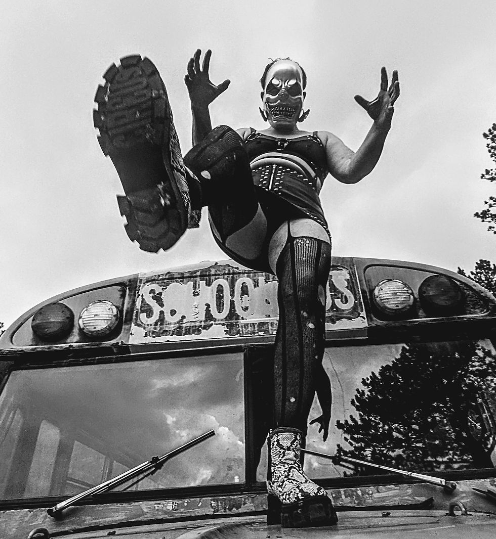 Black-and-white photo of a person wearing a mask, corset, stockings, and platform boots, stepping forward on top of an old bus. Shot from a low angle, their raised foot dominates the foreground, with hands lifted mid-gesture and the bus’s weathered signage visible behind them.