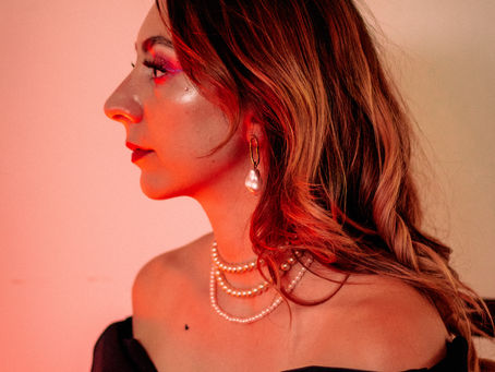 A woman in profile under warm red lighting, wearing an off-shoulder black top, layered pearl necklaces, and a baroque pearl earring, with bold pink and red eye makeup and red lipstick, hair loosely styled over one shoulder.