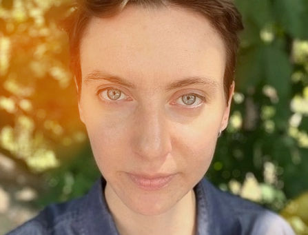 Ryn Zucker has short brown hair and blue eyes and is photographed outdoors in natural light, with green foliage softly blurred in the background. They have a calm expression and are wearing a dark blue shirt