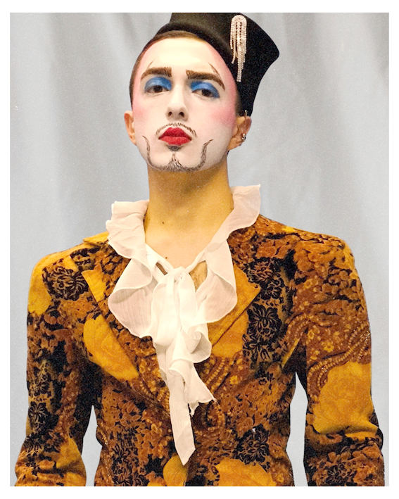 A performer in theatrical stage makeup with a white-painted face, drawn-on beard and moustache, bold blue eyeshadow and red lips. They wear a small tilted black hat adorned with pearl trim, and a richly patterned gold and black brocade jacket with a ruffled white shirt underneath.