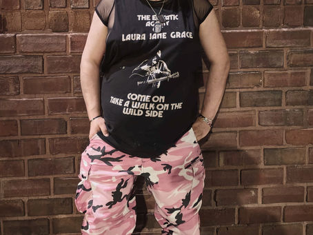 A trans woman in pink camo fatigues, a black leather collar, and Demonia boots stands in front of a brick wall, gazing left with a slight smile and black lipstick.