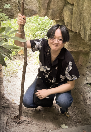 An Asian man with dark hair and glasses crouching in front of an outdoor stone structure. He is holding upright a large stick, taller than he is.