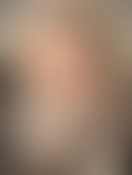 A close-up portrait of a person seated in a car, looking directly at the camera. Their mouth and lower face are covered by a black-and-white patterned keffiyeh, while their brown eyes are in sharp focus. The background is softly blurred, and light falls across their hair and forehead, giving the image a quiet, introspective tone.