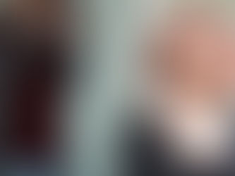 The Vivienne is wearing a red wig, low-cut sparkly top, dramatic eyeshadow and is applying makeup with a brush. John Hyland is standing behind her with a professional camera, taking her photograph.