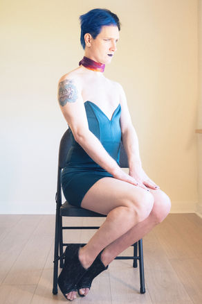 A person with short blue hair and dark lipstick sits sideways on a folding chair, wearing a teal strapless dress and black heeled sandals, with a colourful tattoo visible on their upper arm and a silk scarf at their neck.