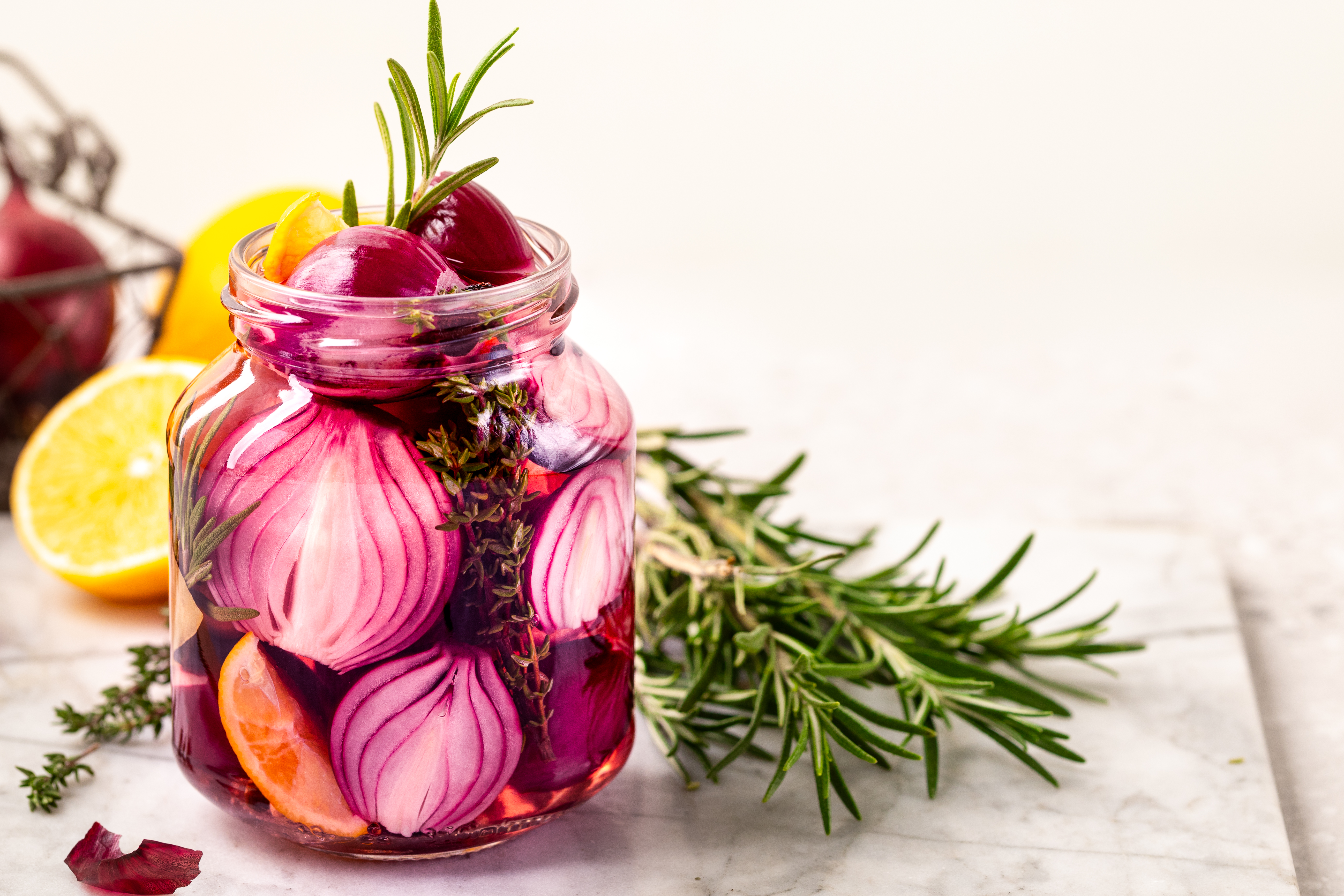 Jar of pickled red onions with rosemary and thyme, set on a marble surface. Halved lemons and fresh herbs in the background.