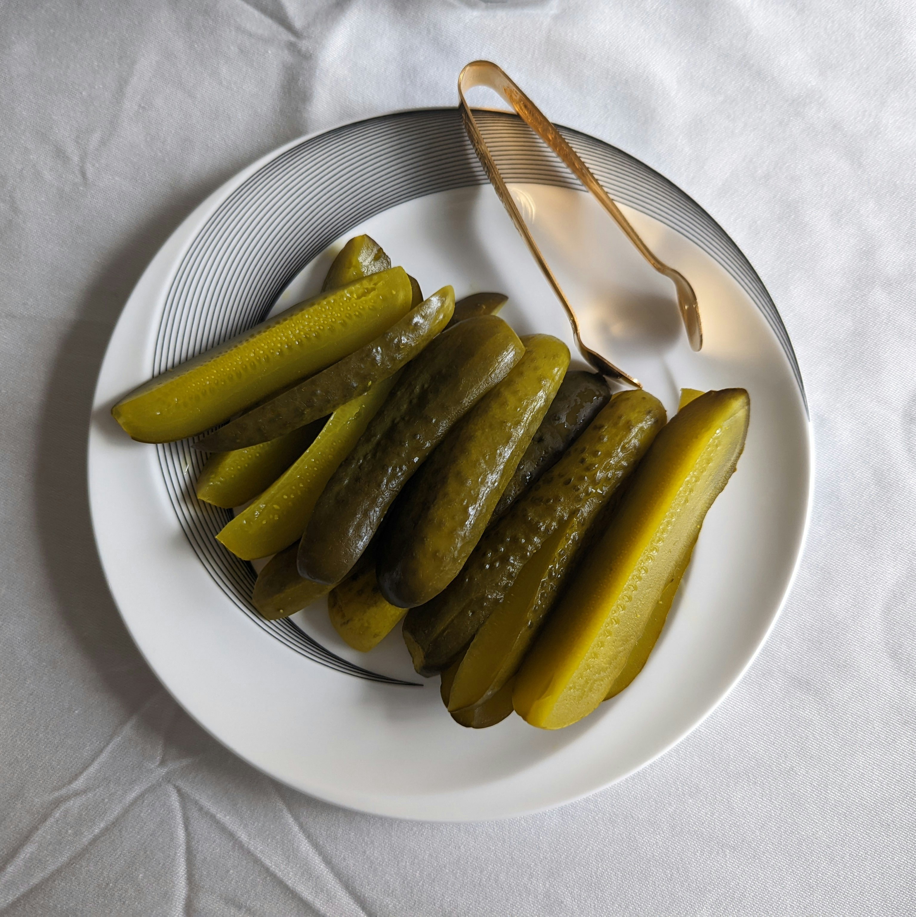 Pickle slices on a white plate with striped rim, accompanied by bronze tongs, set on a white tablecloth.