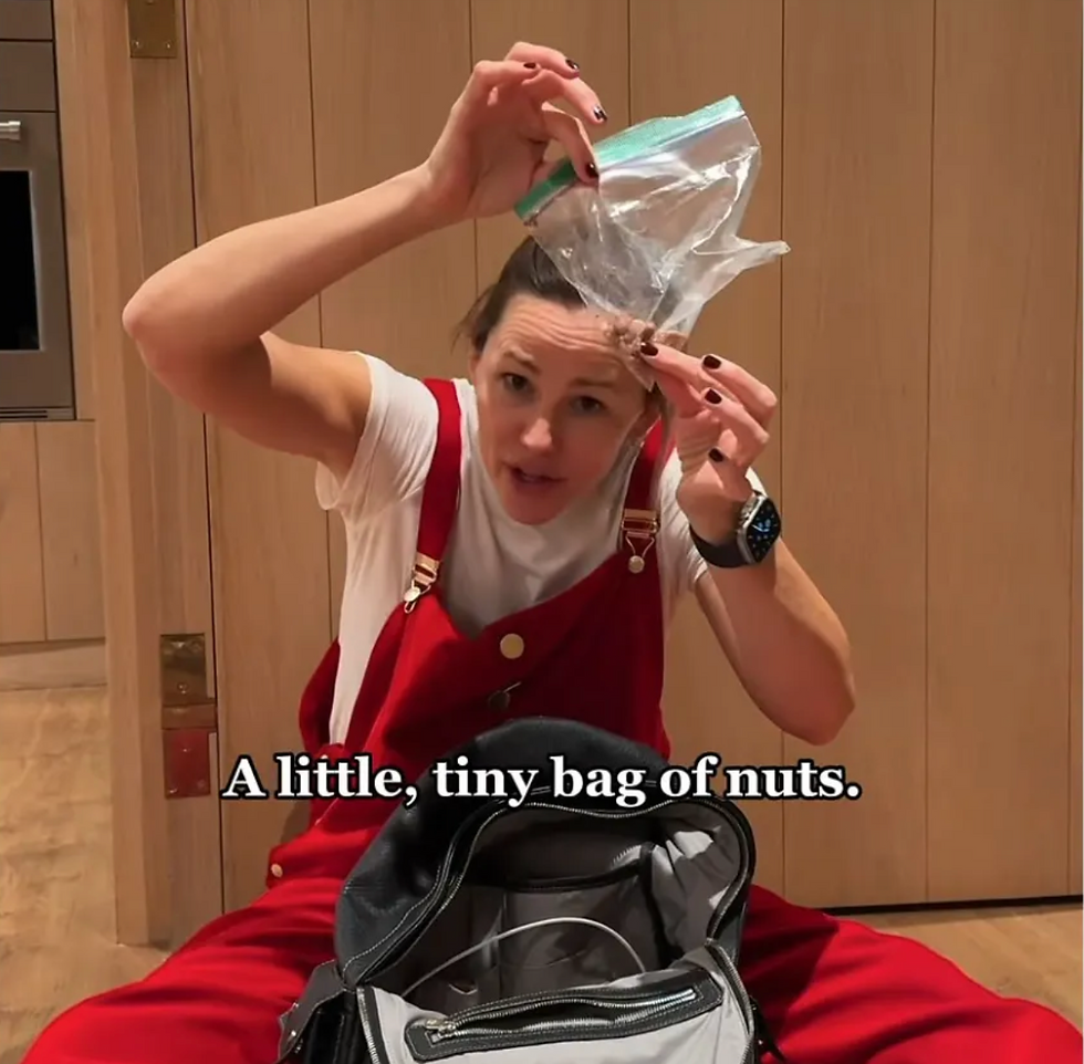 Jennifer Garner in red overalls holds up a tiny plastic bag of nuts in a kitchen; caption reads, A little, tiny bag of nuts.