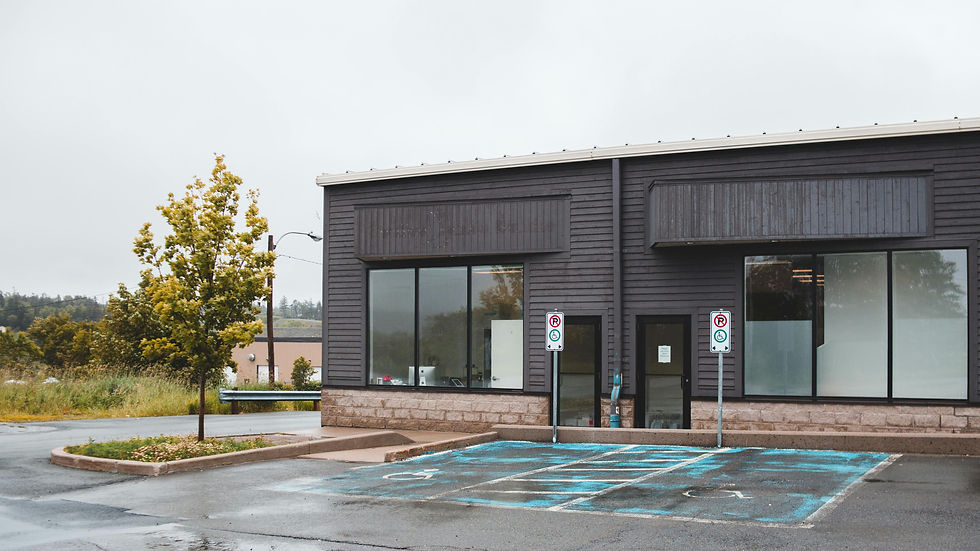 Empty parking lot in front of a grey commercial building with large windows, a tree on the side, and reserved parking signs.