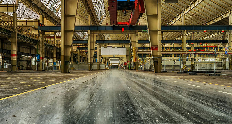 Spacious empty industrial warehouse with high ceilings, yellow beams, and glossy floor. Overhead cranes and yellow lines add structure.