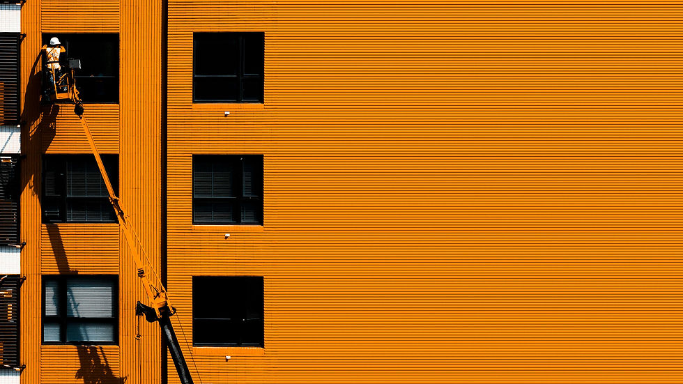 Worker on a lift does maintenance on an orange building with dark windows. Bright, contrasting colors dominate the scene, creating a striking visual.