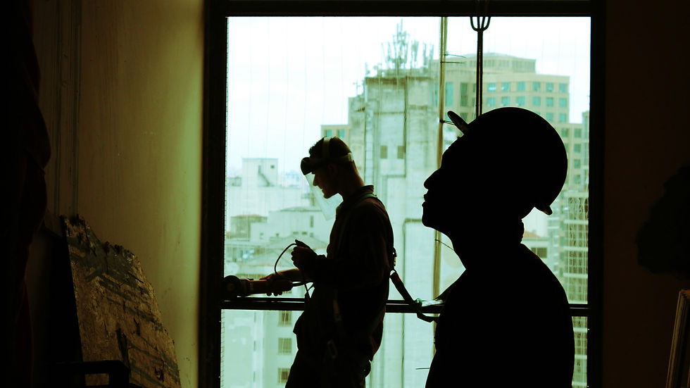 Silhouetted workers in hard hats at a construction site, one adjusting equipment, against a cityscape through a large window. Mood is industrious.
