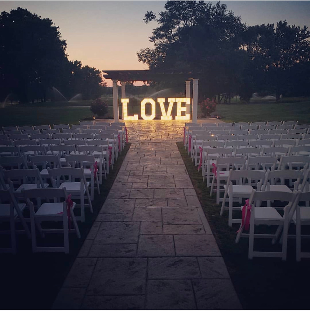 Chairs are set up outside at night for a wedding. At the end of the aisle where the bride and groom would stand are four giant letters with lights inside that spell out the word love.