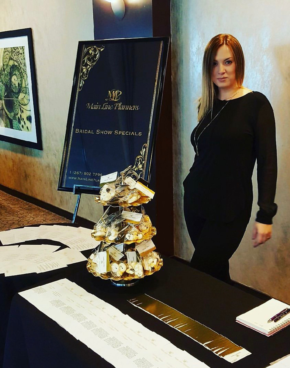 A woman stands behind table at a wedding show. There is a sign that says Main Line Planners next to her. There are business cards and pamphlets on the table.