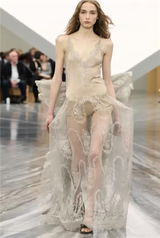 Model walks runway in delicate, sheer beige lace dress. Background features blurred audience on sleek, glossy floor. Elegant mood.