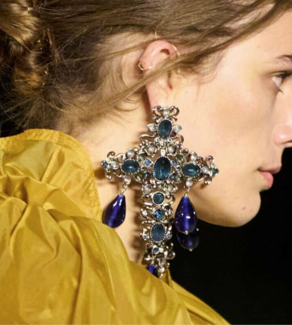 Close-up profile view of a model wearing oversized ornate cross-shaped earrings with blue gemstone details, paired with a yellow textured garment.