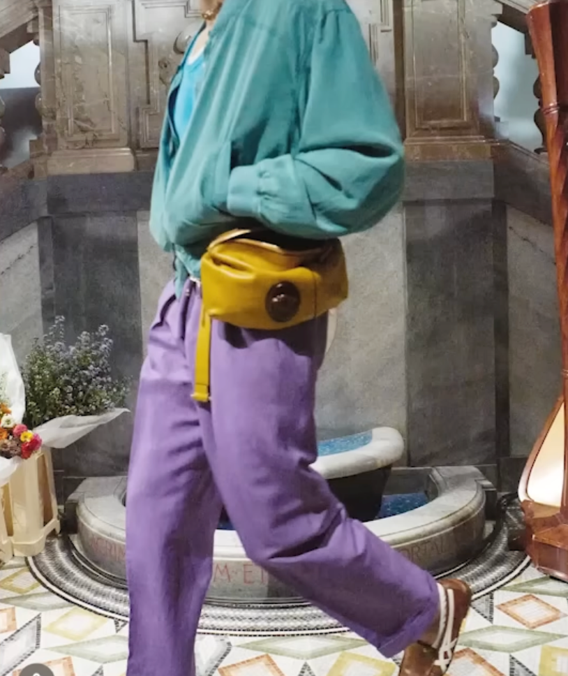 A person in a teal jacket and purple pants walks past a stone fountain. They carry a yellow bag. Background has floral and stone decor.