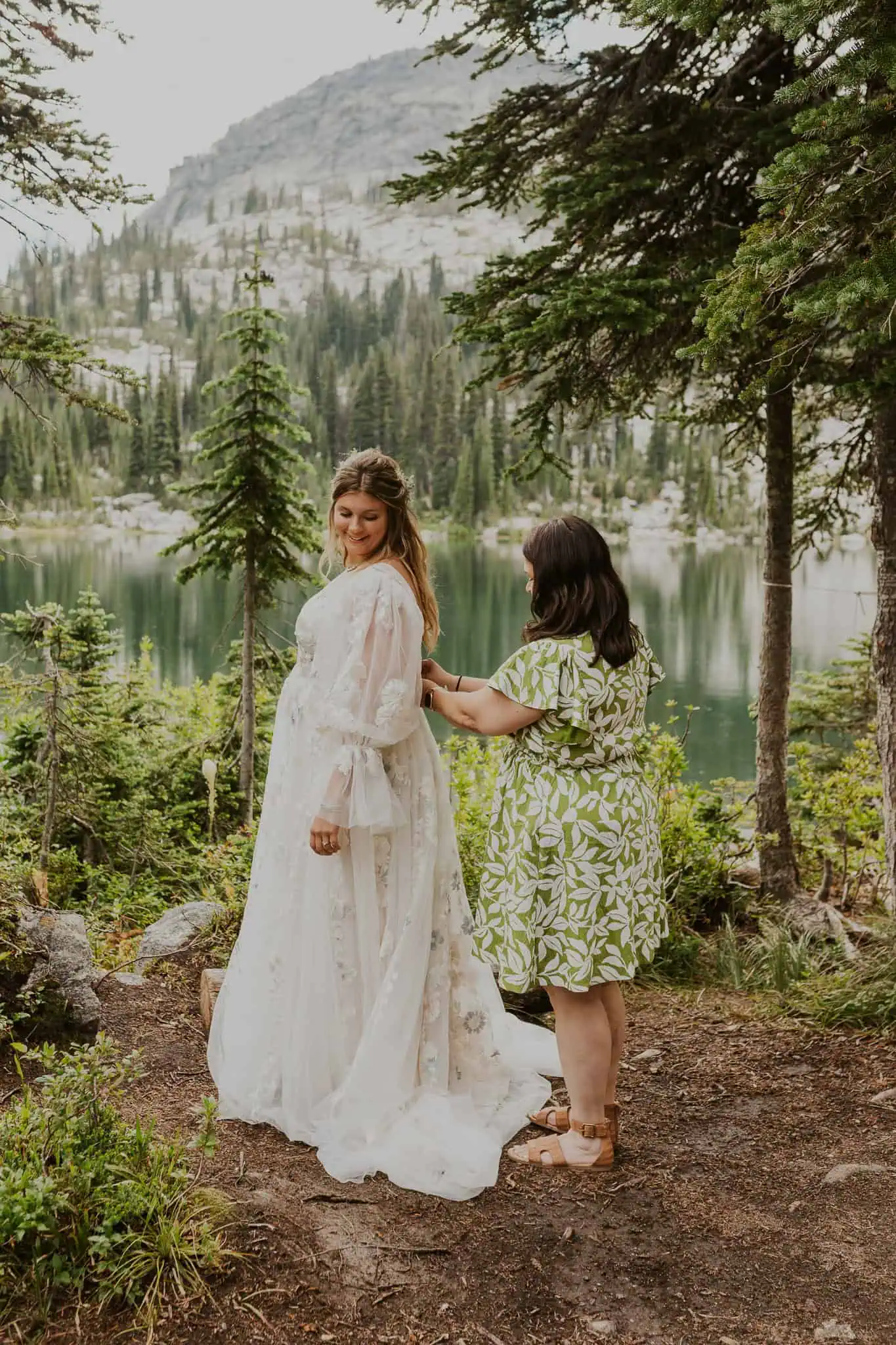bride;s mom helping zip up her dress by an alpine lake
