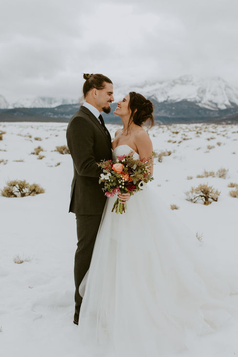 a bride and groom smiling and kissing in front of snow covered mountains