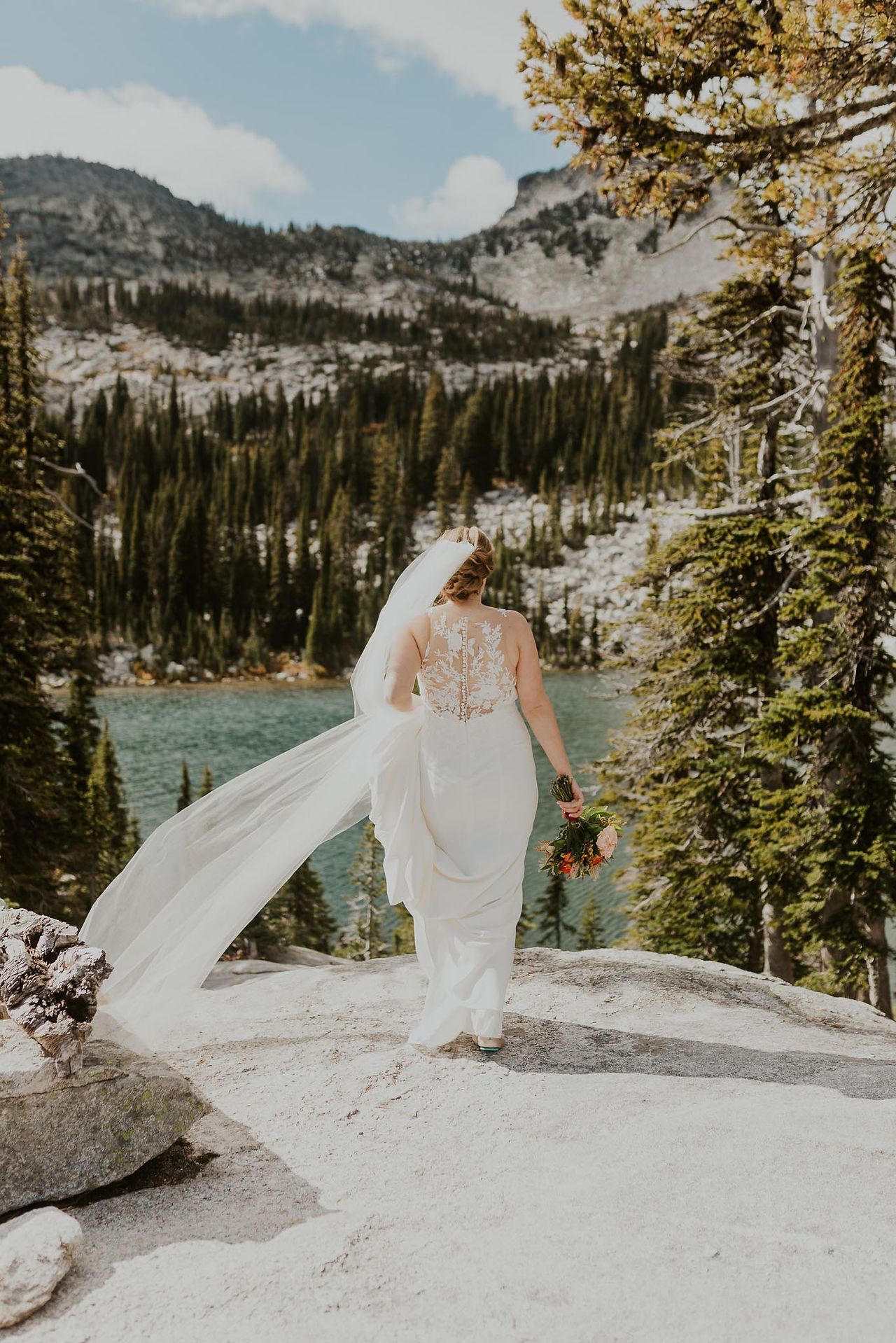 a bride in the mountains holding the train of her dress overlooking a lake