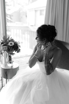 a bride putting in her earrings while sitting in a chair by a window