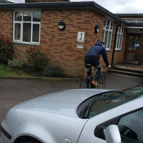 Cyclist in blue jacket rides past a parked silver car toward a brick building with windows. Overcast sky, calm atmosphere.