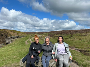 Three people sit smiling on a stone bench in a grassy, hilly landscape under a cloudy blue sky. They wear casual outdoor clothing.