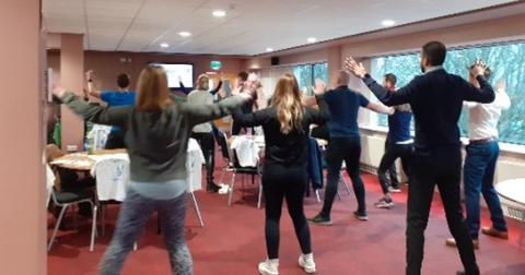 People in a room doing exercises with arms raised. Chairs and tables with white cloths in the background. The mood is active and energetic.