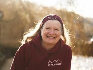 Smiling woman in a maroon hoodie labeled "ST. PAUL STRONG," outdoors by a river. Sunlight and trees create a warm, cheerful atmosphere.