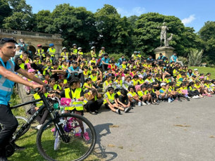 Children sat on the floor next to a man on a bike. They are all wearing high-visibility jacket.