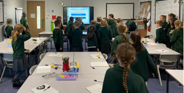 children in school uniforms are in a classroom, gathered round a screen for an activity