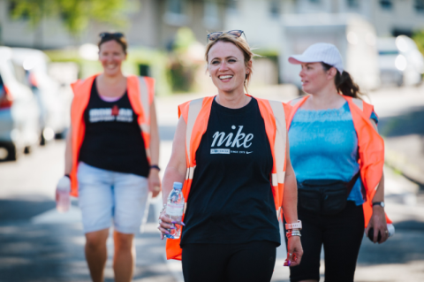three women in high-vis jackets are walking down the road with bottles of water smiling