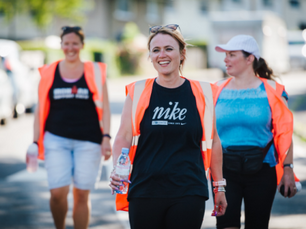 Three women smile and chat as they walk through the street. They all wear an orange vest and hold water bottles.