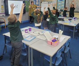 Students in a classroom raise their hands, following a teacher’s lead. A screen displays "10"; bright posters decorate the walls.