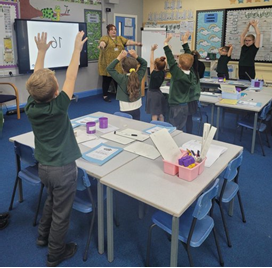 Students in a classroom raise their hands, following a teacher’s lead. A screen displays "10"; bright posters decorate the walls.