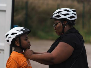 Woman adjusting child's white helmet outdoors, both wearing helmets. Child in orange shirt, woman in black. Overcast day, green background.