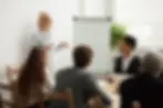 A woman stands by a flip chart, presenting to a group of four seated colleagues in a meeting room. T