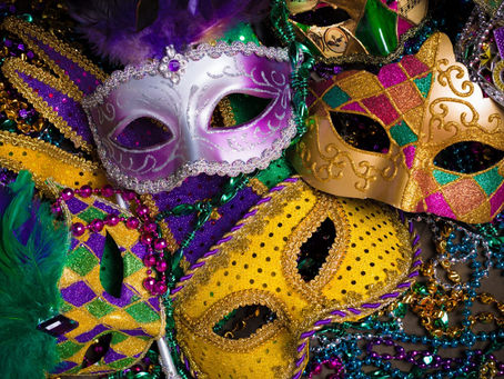 Pile of glittery Mardi Gras masks in purple, gold and green, with beads and feathers.